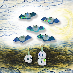 May include: A whimsical illustration features several bats with blue and iridescent wings, and two skull-shaped candle holders. The background includes a yellow crescent moon, blue clouds, and a dark, textured landscape.