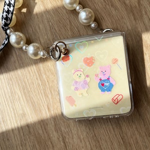 May include: A clear plastic phone case with a light yellow background and colorful cartoon stickers of bears, hearts, and candy. The case is attached to a pearl and black and white checkered ribbon.