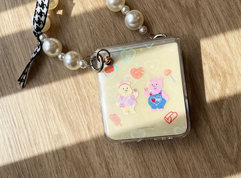 May include: A clear plastic phone case with a light yellow background and colorful cartoon stickers of bears, hearts, and candy. The case is attached to a pearl and black and white checkered ribbon.