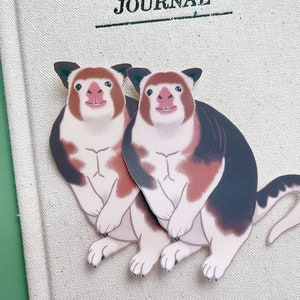 May include: Two cartoon red-brown and black animal stickers with white markings. The animals are standing on their hind legs with their front paws raised. They have white faces with pink noses and black eyes.