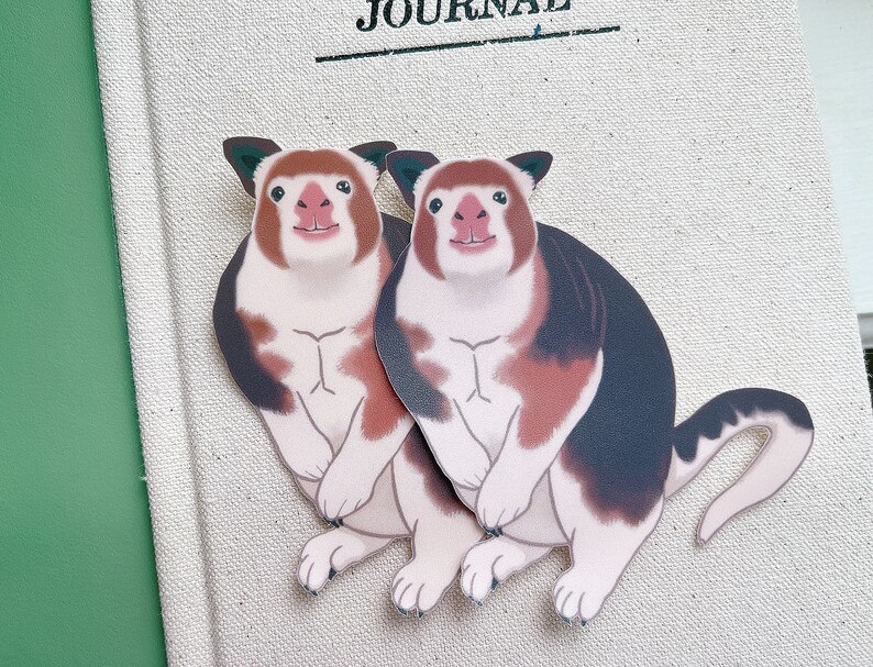 May include: Two cartoon red-brown and black animal stickers with white markings. The animals are standing on their hind legs with their front paws raised. They have white faces with pink noses and black eyes.