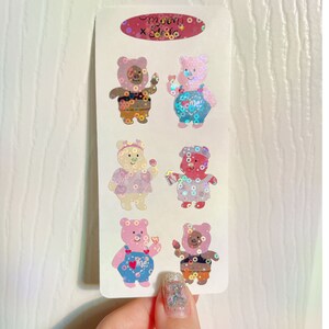 May include: A sheet of six holographic stickers featuring cartoon bears wearing different outfits. The bears are pink, brown, and white. The stickers are on a white background.