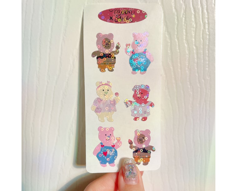 May include: A sheet of six holographic stickers featuring cartoon bears wearing different outfits. The bears are pink, brown, and white. The stickers are on a white background.