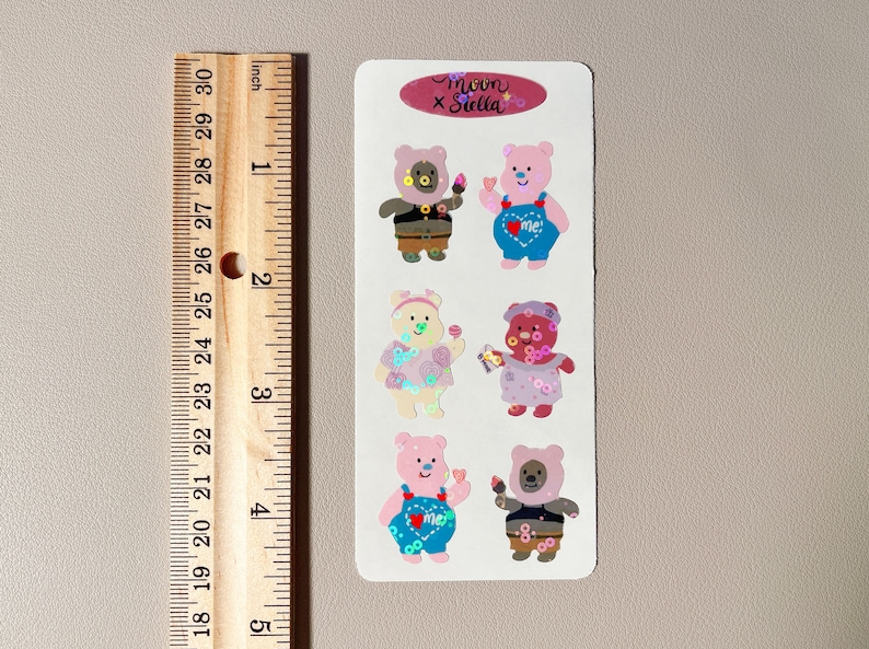 May include: A sheet of stickers featuring six adorable cartoon bears wearing different outfits. The bears are pink, brown, and white with colorful details. The stickers are labeled "moon x stella".