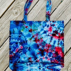 May include: A tie-dye tote bag with a vibrant pattern of blue, red, and white. The bag has matching tie-dye handles and is made of a sturdy fabric. The bag is sitting on a wooden surface.
