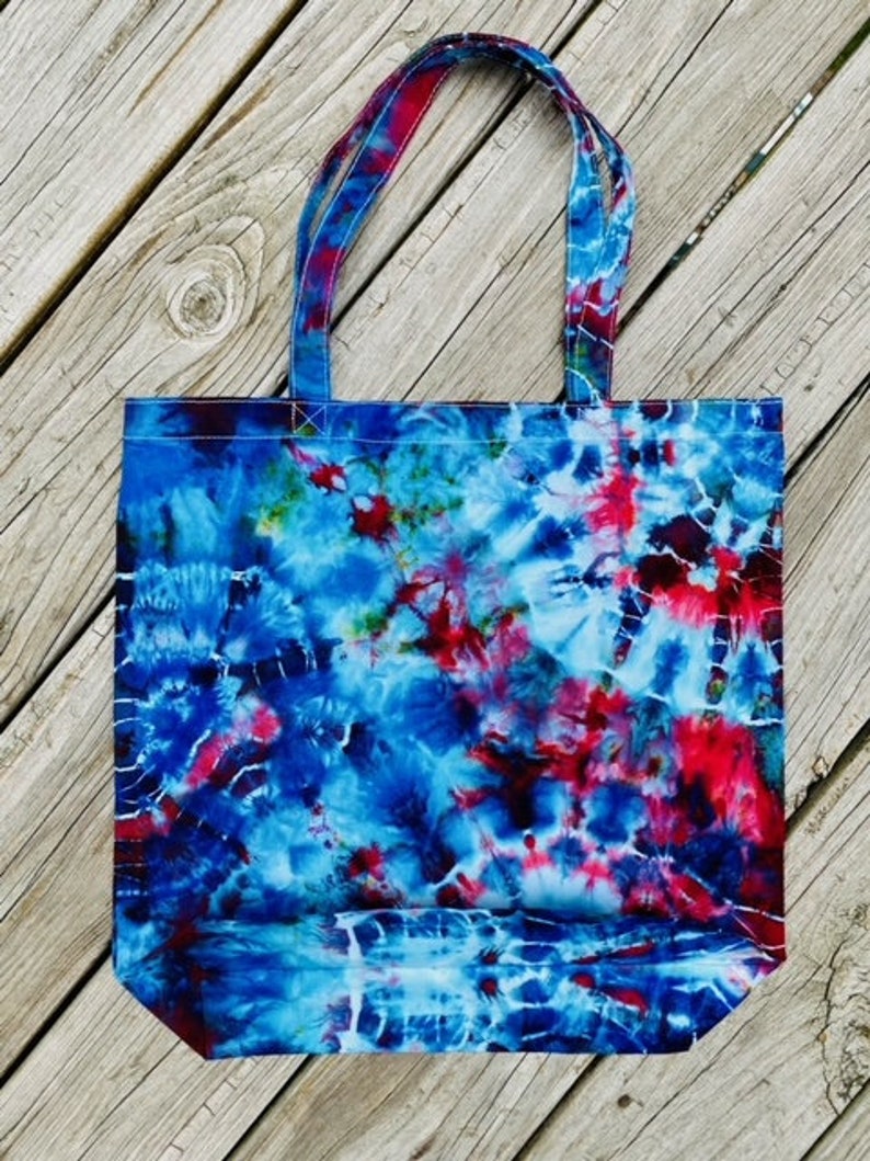 May include: A tie-dye tote bag with a vibrant pattern of blue, red, and white. The bag has matching tie-dye handles and is made of a sturdy fabric. The bag is sitting on a wooden surface.