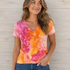 May include: A tie-dye V-neck t-shirt in pink and orange hues. The short-sleeved top is made of a soft fabric and is worn with blue jeans. The shirt's design features a blend of bright colours, creating a visually appealing pattern.