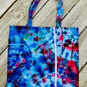 May include: A tie-dye tote bag with blue, red, and white swirls. The bag has matching handles and a white measuring tape is placed vertically against the bag. The bag is made of fabric and is sitting on a wooden surface.