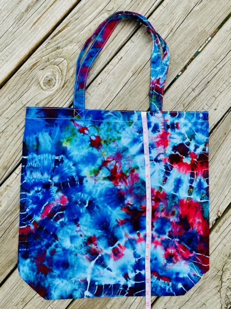 May include: A tie-dye tote bag with blue, red, and white swirls. The bag has matching handles and a white measuring tape is placed vertically against the bag. The bag is made of fabric and is sitting on a wooden surface.