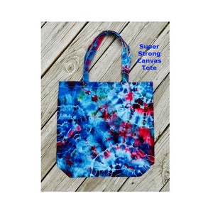 May include: A tie-dye canvas tote bag in shades of blue, red, and white. The bag features strong handles and the text "Super Strong Canvas Tote". This tote bag is designed for carrying items.