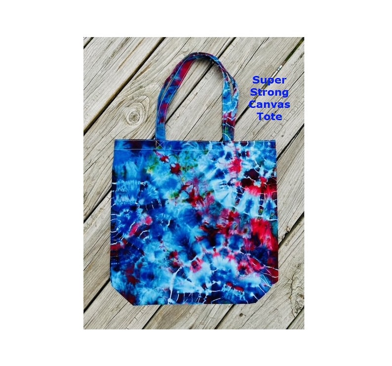 May include: A tie-dye canvas tote bag in shades of blue, red, and white. The bag features strong handles and the text "Super Strong Canvas Tote". This tote bag is designed for carrying items.