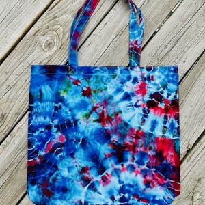 May include: A fabric tote bag featuring a tie-dye design in shades of blue, red, and white. The bag has two handles and a rectangular shape. The tie-dye pattern is spread across the entire surface, creating a colourful accessory.