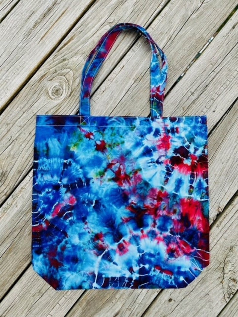 May include: A fabric tote bag featuring a tie-dye design in shades of blue, red, and white. The bag has two handles and a rectangular shape. The tie-dye pattern is spread across the entire surface, creating a colourful accessory.