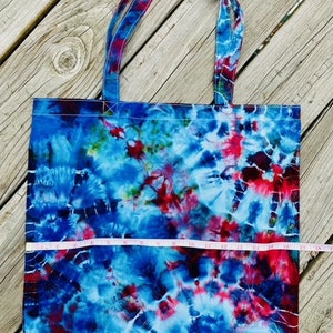 May include: A tie-dye tote bag with blue, red, and white swirls. The bag has two handles and a rectangular shape. The bag is approximately 38 cm wide.