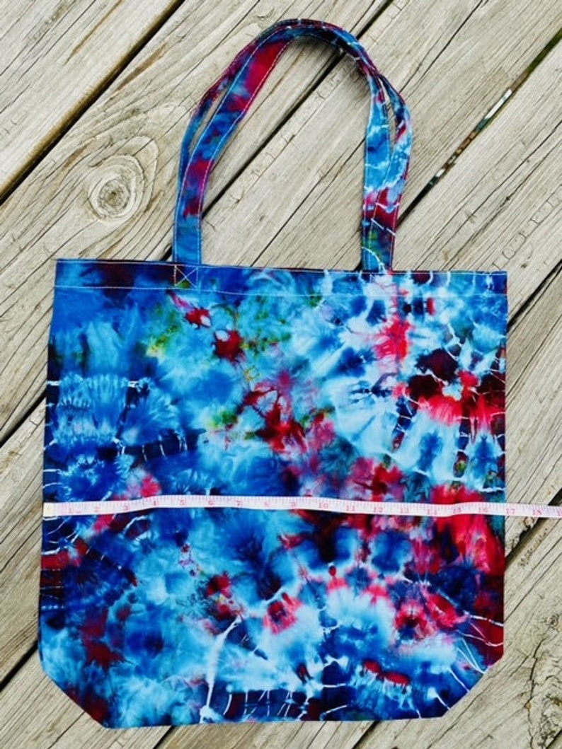 May include: A tie-dye tote bag with blue, red, and white swirls. The bag has two handles and a rectangular shape. The bag is approximately 38 cm wide.