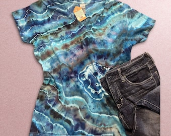 May include: A blue and green tie-dye v-neck t-shirt with short sleeves. The shirt is lying on a pink surface with a pair of dark blue jeans.