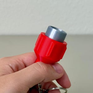 Könnte beinhalten: Ein rotes und graues Schlüsselanhänger-Zubehör. Der rote Teil ist zylindrisch mit einem geriffelten Design, und der graue Abschnitt ist ein kleinerer Zylinder. Ein silberner Schlüsselring ist befestigt.