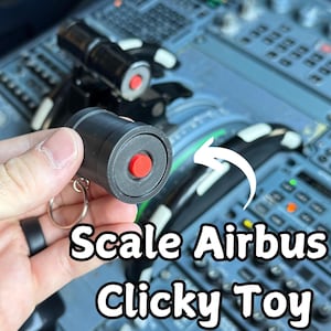 May include: A black and red scale Airbus clicky toy with a keyring. The toy is cylindrical with a red button on the end. The background shows an aircraft cockpit.