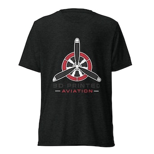 May include: A dark heather gray short-sleeve t-shirt featuring a graphic of a three-blade airplane propeller centered over a red and white circular emblem. Below the emblem, the text reads 3D PRINTED AVIATION in gray and red lettering.