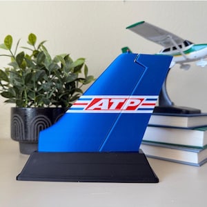 May include: Blue and white airplane tail fin with the word "ATP" in red letters. The tail fin is on a black base.