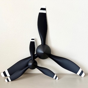 May include: Two black airplane propellers with white-striped tips are displayed against a neutral background. The propellers are of different sizes, with three blades each, and are mounted on black hubs. The blades have a sleek, curved design.