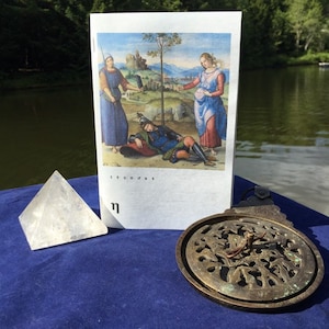 May include: A clear quartz pyramid, a vintage brass astrolabe, and a postcard featuring a Renaissance painting with figures in blue and red robes. The items are arranged on a blue surface near a body of water.