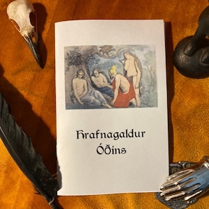 May include: A white booklet with the title "Krafnagaldur Óðins" and a color illustration of figures in a forest setting. The booklet is on an orange surface, with a feather quill, a bird skull, and a hand-shaped metal object.