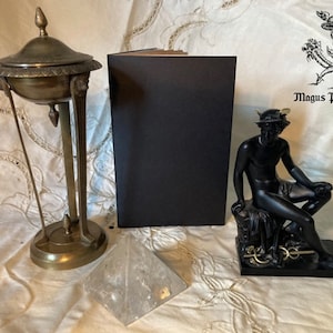 May include: An antique brass incense burner, a black statue of a seated figure, a clear quartz pyramid, and a black book are arranged on a patterned cloth. The statue is of a man wearing a winged helmet. The text "Magus Publishing" is visible.