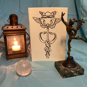 May include: A still life arrangement featuring a lit candle inside a vintage metal lantern, a clear quartz crystal ball, a drawing of a caduceus symbol, and a bronze statue of a figure with a staff. The items are arranged on a blue surface.