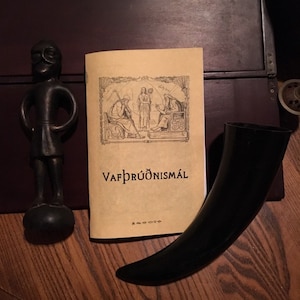 May include: An arrangement of Viking-themed items. A black figurine stands next to a book titled "Vafþrúðnismál" with illustrations. A black drinking horn is also present. The items are on a wooden surface.