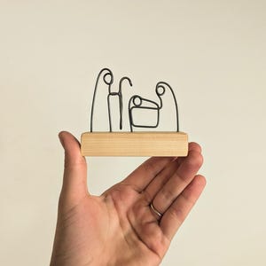 May include: A wire sculpture of the nativity scene on a wooden base. The sculpture is made of black wire and depicts Mary, Joseph, and the baby Jesus.