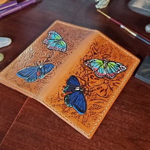 Cartera Mariposa que combina estilos tradicionales de cuero y tallado de figuras con pintura colorida en una pieza hecha a mano y funcional.