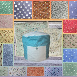 May include: A teal and white fabric ottoman with a white pocket and a button. The ottoman is surrounded by fabric swatches in various floral patterns and colors, including green, blue, and red. The background is a light wood.