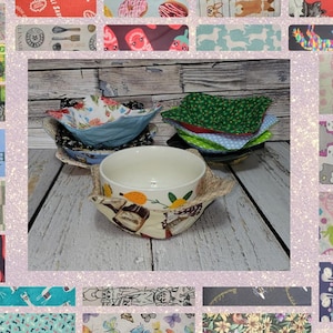 May include: A set of four bowl cozies made from fabric with various patterns. The cozies are folded and stacked on top of each other. A white ceramic bowl with a floral pattern is sitting on top of the cozies.