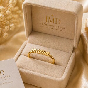 Peut inclure: Une bague dorée avec le mot "mama" dans un écrin à bijoux. La bague est dans un écrin en velours beige avec le logo "JMD Sterling Silver". Une boîte cadeau avec un nœud et une carte avec le même logo sont également visibles.