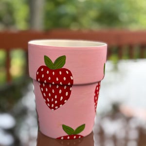 May include: A pink painted flower pot with a repeating strawberry design. The strawberries are red with white seeds and green leaves. The pot is made of ceramic and is suitable for indoor or outdoor use.