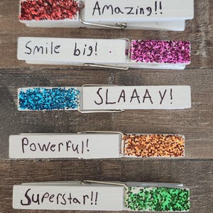 May include: Five wooden clothespins with glitter on the top half. Each clothespin has a different color glitter and a handwritten message. The messages are "Amazing!!", "Smile big!", "SLAAAAY!", "Powerful!", and "Superstar!!".