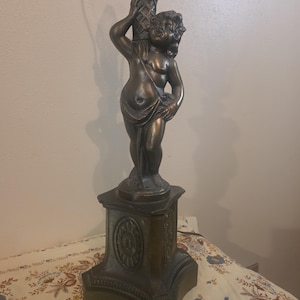 Puede incluir: Lámpara de mesa de color bronce con una figura de querubín sosteniendo un elemento decorativo. La lámpara tiene una base detallada con detalles florales y un portalámparas de latón. El querubín está cubierto con tela. Es una lámpara decorativa.