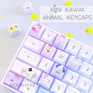 May include: A white keyboard with colorful keycaps featuring kawaii animal designs. The keycaps are shaped like a dog, cat, bunny, and other animals. The keyboard has a rainbow gradient effect. The text "MINI KAWAII ANIMAL KEYCAPS" is displayed above the keyboard. The text "MOON N' KEYS" is displayed below the keyboard.
