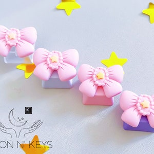 May include: Pink bow-shaped keycaps with a yellow star center, resting on light purple and clear bases. Yellow star decorations are scattered around the keycaps. The Moon N' Keys logo is in the lower left corner.