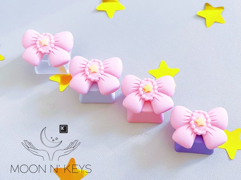 May include: Pink bow-shaped keycaps with a yellow star center, resting on light purple and clear bases. Yellow star decorations are scattered around the keycaps. The Moon N' Keys logo is in the lower left corner.