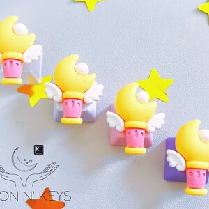 May include: Four yellow moon-shaped wands with white wings and pink bases are displayed. Each wand is attached to a square, with yellow star accents. The Moon N' Keys logo is visible in the lower left corner.