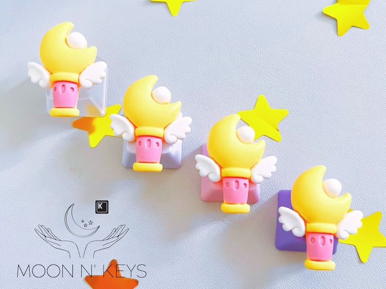 May include: Four yellow moon-shaped wands with white wings and pink bases are displayed. Each wand is attached to a square, with yellow star accents. The Moon N' Keys logo is visible in the lower left corner.