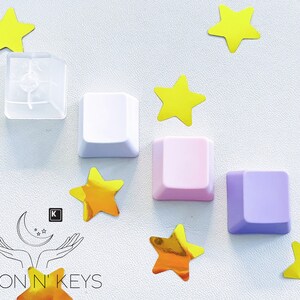May include: A collection of colorful keyboard keycaps in white, pink, and purple, arranged with yellow star-shaped confetti. One keycap is transparent. The Moon N' Keys logo is in the lower left corner.