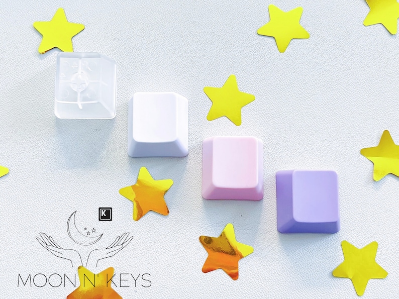 May include: A collection of colorful keyboard keycaps in white, pink, and purple, arranged with yellow star-shaped confetti. One keycap is transparent. The Moon N' Keys logo is in the lower left corner.