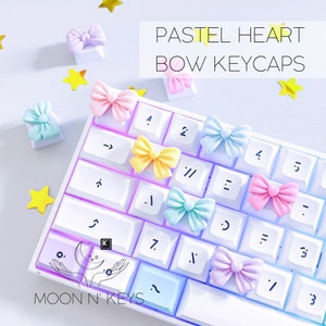 May include: A white keyboard with pastel colored bow keycaps. The keycaps are in the colors pink, blue, yellow, and purple. The keyboard has a rainbow light effect. The text "PASTEL HEART BOW KEYCAPS" is at the top of the image. The text "MOON N' KEYS" is at the bottom of the image.