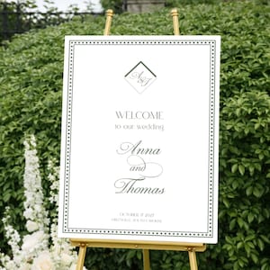 May include: A wedding welcome sign on a gold easel. The white sign has a black dotted border and reads "Welcome to our wedding" with the names "Anna and Thomas" in elegant script. The sign is set against a backdrop of greenery and flowers.
