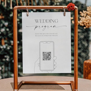 May include: A copper-colored metal frame holds a white sign with the words "WEDDING program" in elegant script. The sign includes a QR code and instructions to scan for details. A small red flower adorns the top of the frame.