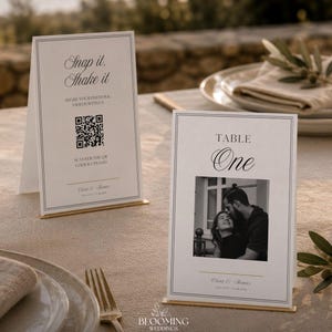 May include: Two white rectangular table signs with thin black borders and gold-toned metal stands. One sign displays the text Snap it, Shake it, a QR code, and the names Chloe & Thomas. The other sign reads TABLE One with a black and white photo.