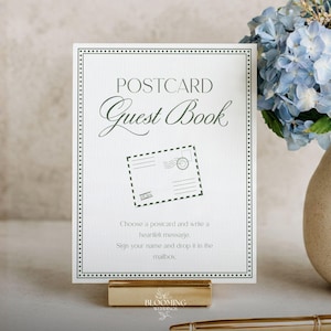 May include: A white postcard guest book with the text "POSTCARD Guest Book" and instructions. The sign is in a gold holder, with a vase of blue and white flowers in the background.